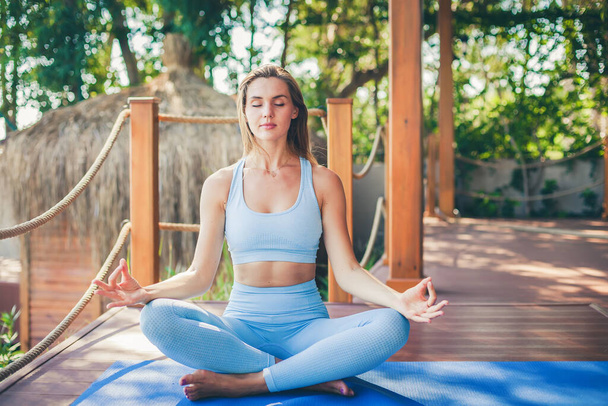Beautiful relaxing woman with closed eyes sitting in a lotus position while doing yoga in a tropical resort. Healthy lifestyle and self body care. - Zdjęcie, obraz