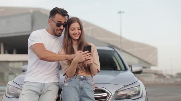 Mężczyzna i dziewczyna stoją w pobliżu samochodu. Uśmiechają się i oglądają coś na swoim smartfonie. Rozmawiają ze sobą. 4K - Materiał filmowy, wideo