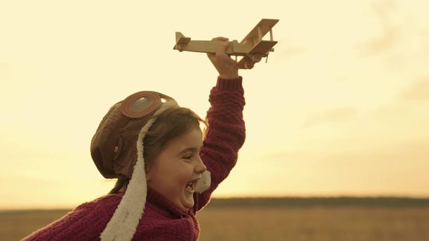 Το μικρό χαρούμενο κορίτσι τρέχει με ένα ξύλινο αεροπλάνο στο χέρι στο ηλιοβασίλεμα στον ουρανό και γελάει, τρέχει παιδί παίζει το παιχνίδι που πετάει πάνω από το έδαφος, τα παιδιά τρέχουν έξω, το παιδί πετάει όνειρο σε ταξίδια διακοπών, το μωρό Σαββατοκύριακο - Φωτογραφία, εικόνα
