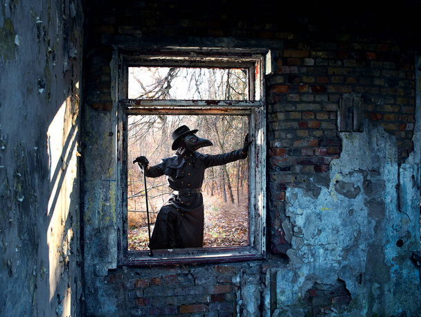 A man in a plague doctor costume in a mask with a beak in the window opening of a destroyed house. - Photo, Image