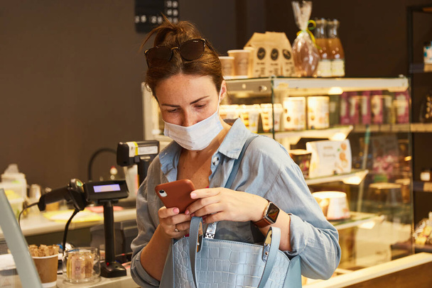 Woman in mask standing in coffee shop and waiting for order. Using her phone. New normal - Photo, Image