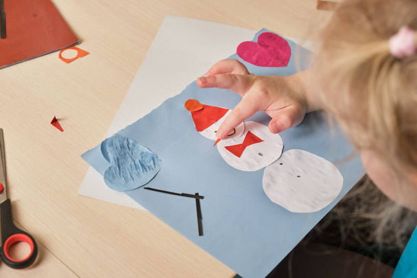 a preschooler girl in a blue T-shirt makes an applique while sitting at the table, a paper snowman hauled on colored paper, children's creativity in winter, atopic dermatitis on the hands of a child - Foto, Imagen
