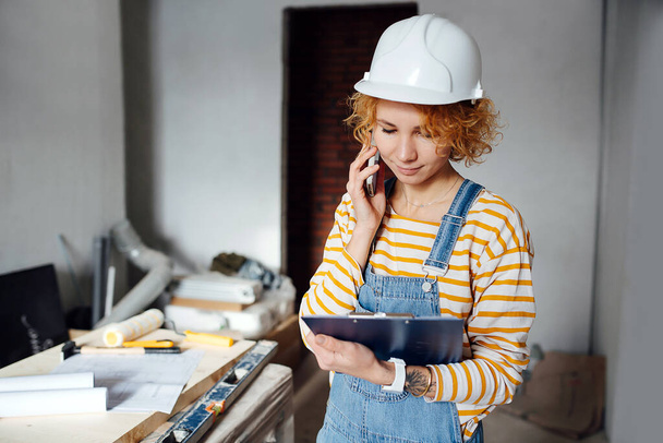 Subtly smiling female construction engineer in a white helmet looking down at the clipboard, speaking on the phone. In a room inside a building under construction. - Photo, image