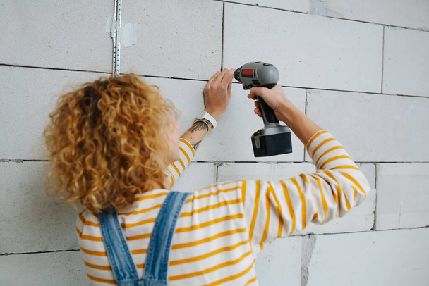 Бачачи вигляд жінки будівельника буріння сірої цегляної стіни з електричною викруткою
. - Фото, зображення
