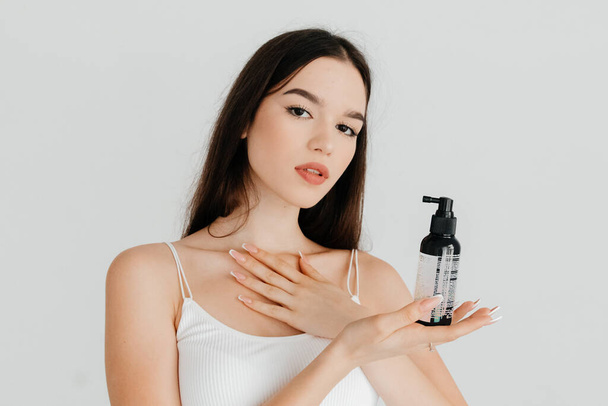 Studio portrait of a pretty young woman with long straight hair and beautiful makeup in a white top posing, holding a bottle of hair product, on a white background - Foto, Imagen