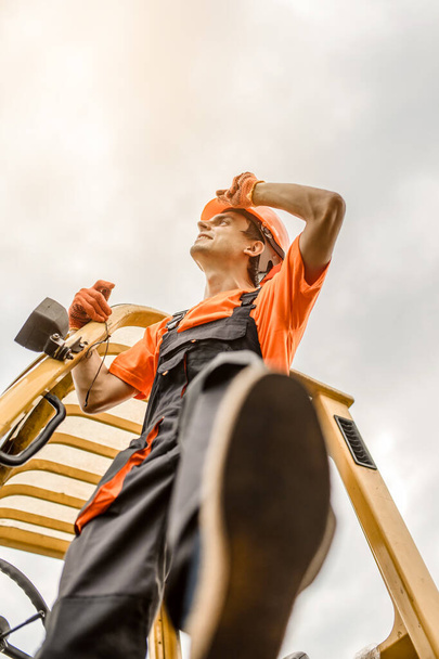 Niskopunktowe ujęcie młodego wykonawcy w kombinezonie i pomarańczowym hełmie ochronnym wydostaje się z maszyny ładującej na placu budowy - Zdjęcie, obraz