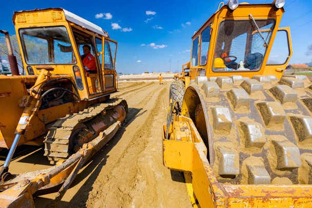 Rodillo de carretera con espigas y bulldozer están compactando, nivelando la arena para la fundación de carreteras en el sitio de construcción. - Foto, imagen