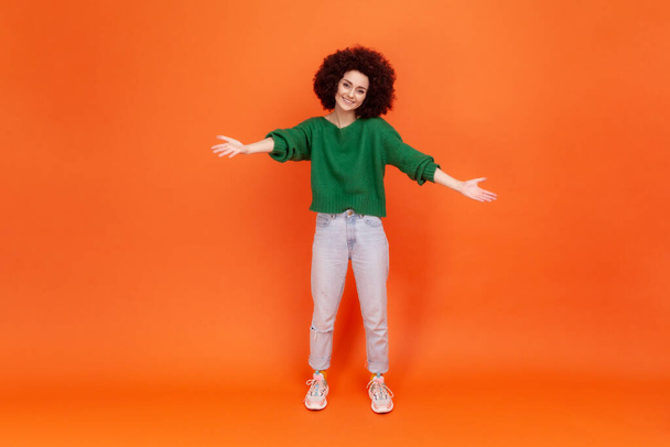 Full length of woman with Afro hairstyle sharing opening hands looking at camera with kind smile, greeting and regaling, happy glad to see you. Indoor studio shot isolated on orange background. - Fotó, kép