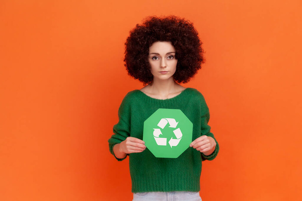Serious woman with Afro hairstyle wearing green casual style sweater holding in hands green recycling sing, ecology concept, looking at camera. Indoor studio shot isolated on orange background. - Photo, Image