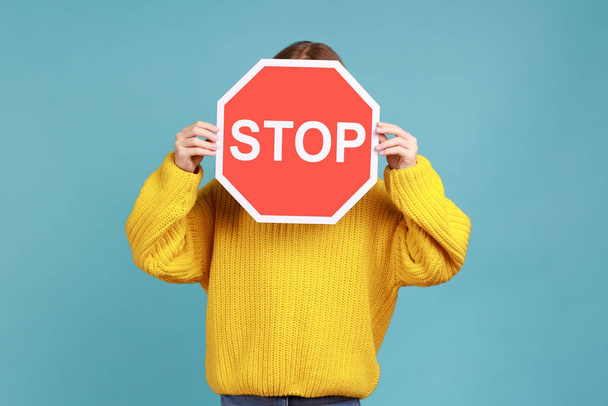 Portrait of unknown little kid covers face with Stop symbol, anonymous child holds red traffic sign, wearing yellow casual style sweater. Indoor studio shot isolated on blue background. - Foto, Bild