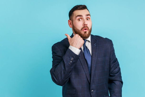 Astonished bearded man wearing dark suit pointing finger aside, showing copy space for commercial text, blank wall with idea presentation. Indoor studio shot isolated on blue background. - Foto, imagen