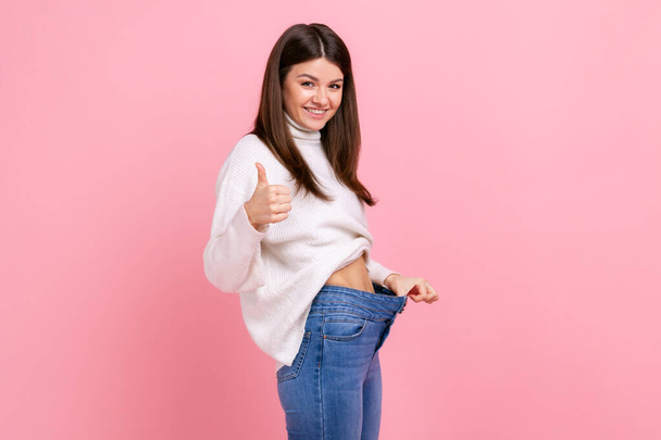 Side view portrait of happy beautiful woman showing thumb up and her slim waist and big trousers, wearing white casual style sweater. Indoor studio shot isolated on pink background. - 写真・画像