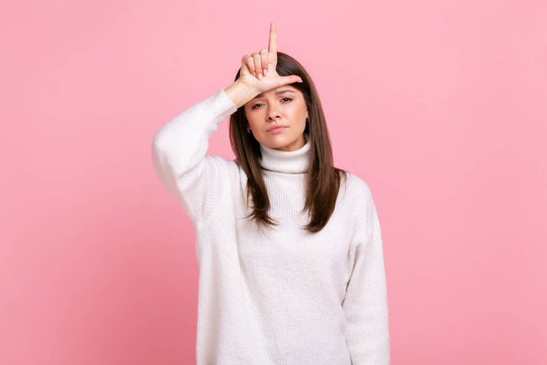 Depressed woman showing loser gesture, L finger sign on forehead, upset about dismissal, unlucky day, wearing white casual style sweater. Indoor studio shot isolated on pink background. - 写真・画像