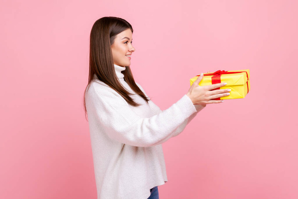 Side view portrait of young adult brunette female holding out yellow wrapped gift box, giving present, wearing white casual style sweater. Indoor studio shot isolated on pink background. - Foto, Imagem