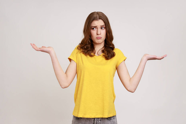 I don't know, who cares. Portrait of confused clueless attractive teenager girl in yellow t-shirt shrugging shoulders, making no idea gesture, whatever. Indoor studio shot isolated on gray background. - Foto, Imagen