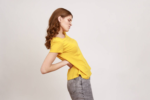 Side view portrait of exhausted unhealthy teen girl in yellow T-shirt touching aching back, suffering lower lumbar discomfort, injured spine disk. Indoor studio shot isolated on gray background. - 写真・画像