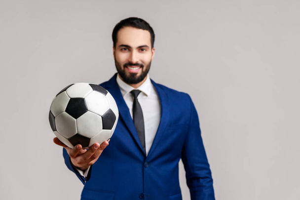 Happy attractive bearded businessman holding out soccer ball on his hand with smiling optimistic expression, wearing official style suit. Indoor studio shot isolated on gray background. - Fotografie, Obrázek