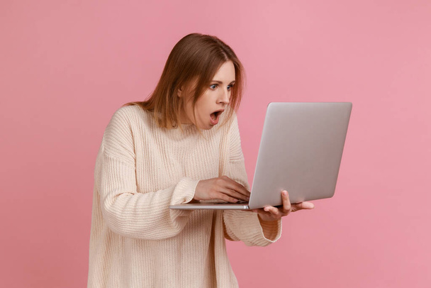 Portrait of amazed shocked young adult blond woman working on laptop compute, looking at display with open mouth, wearing white sweater. Indoor studio shot isolated on pink background. - Foto, imagen