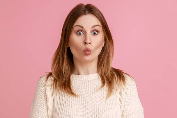 Portrait of blond woman showing fish face grimace with pout lips, fooling around, making ridiculous childish comical grimace, wearing white sweater. Indoor studio shot isolated on pink background. - Photo, image