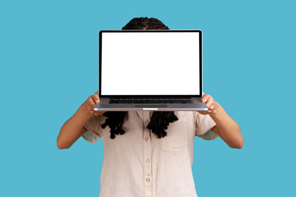 Portrait of unrecognizable woman hiding her face behind laptop with blank display for advertisement or promotional text, wearing white shirt. Indoor studio shot isolated on blue background. - Fotoğraf, Görsel