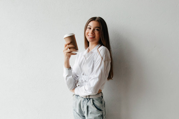 Young brunette woman holding disposable coffee cup and smiling, looking at the camera - Foto, afbeelding