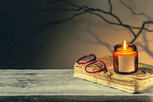 Vintage background with an old antique book, glasses and candle in a glass jar burning at night. Tree brunches and shadows make spooky dark academia atmosphere. Cozy fall season reading time - Photo, Image