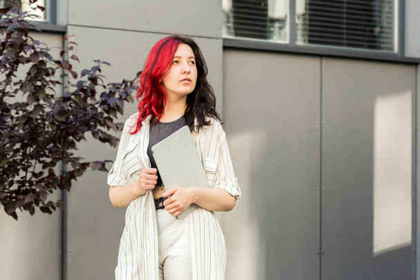 Portrait of a female student or businesswoman with creative red and black hair in smart casual clothing using digital tablet outdoors near the office building - Foto, immagini