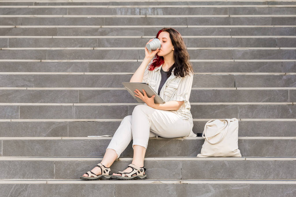 Beautiful White young business woman holding Digital Tablet outdoors sitting on the stairs during her coffee break. Drinking coffee from reusable collapsible cup, Sustainable lifestyle concept - Фото, изображение