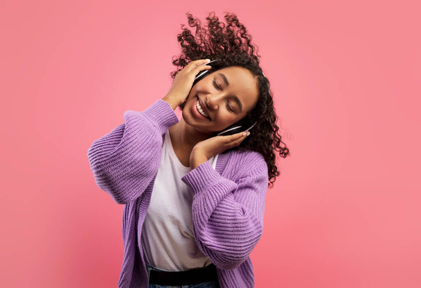 Carefree African American lady listening to music in wireless headphones, dancing with closed eyes on pink background - Fotografie, Obrázek