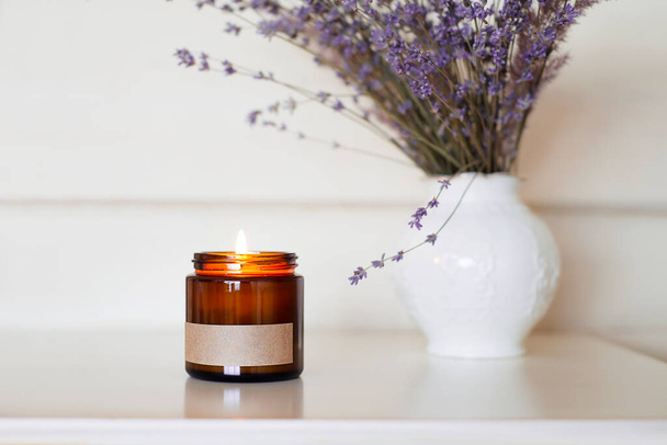 Glass red-brown jar a burning candle on the on a nightstand with lavender flowers. Home comfort concept with aromain provence style. Label for inscription. - Photo, Image