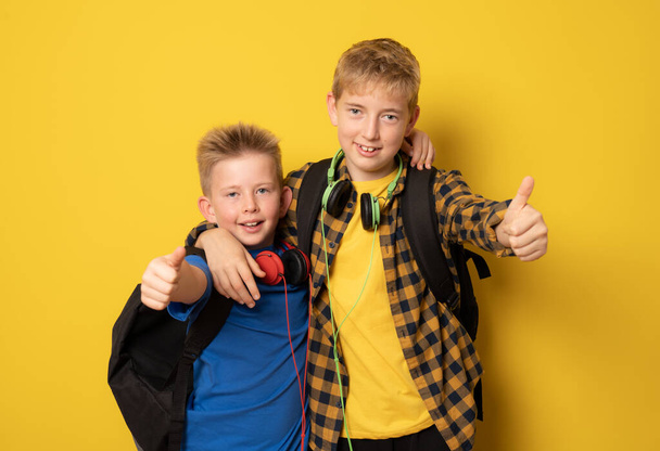 2人の学校の男の子がヘッドフォンをつけて黄色の背景に親指を上げ. - 写真・画像