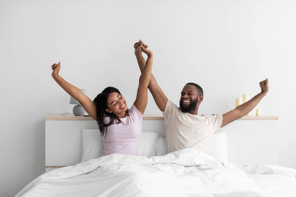 Satisfied young african american guy and lady raise hands, stretching body on bed in bedroom interior - Fotografie, Obrázek