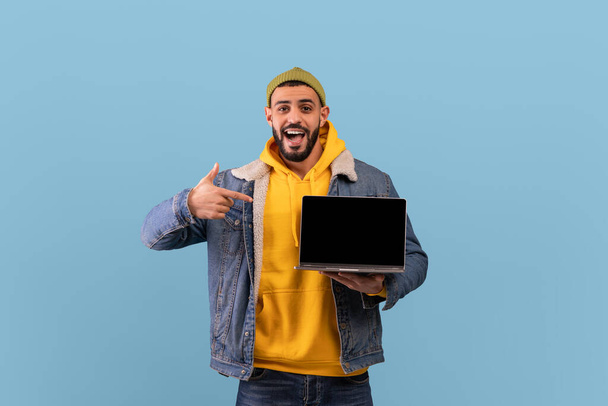 Overjoyed hipster arab guy pointing at laptop computer with blank screen, posing over blue studio background, mockup - Photo, Image