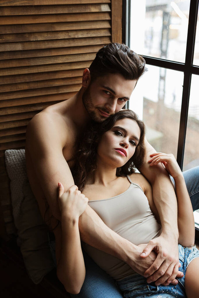 portrait of a charming young couple at home. man is embracing his girlfriend near window. Two people relaxing together - Foto, Imagen