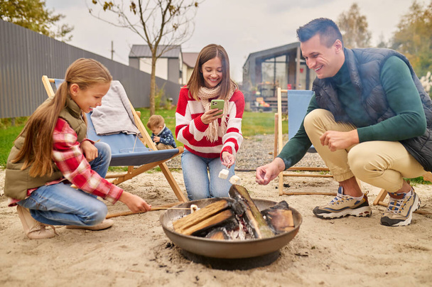 Family holding marshllow on stick over campfire in yard - 写真・画像