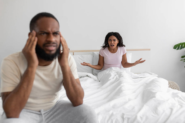 Δυσαρεστημένη νεαρή μαύρη γυναίκα ορκίζεται και φωνάζει στον θλιμμένο προσβεβλημένο σύζυγο στο κρεβάτι στο εσωτερικό της λευκής κρεβατοκάμαρας - Φωτογραφία, εικόνα