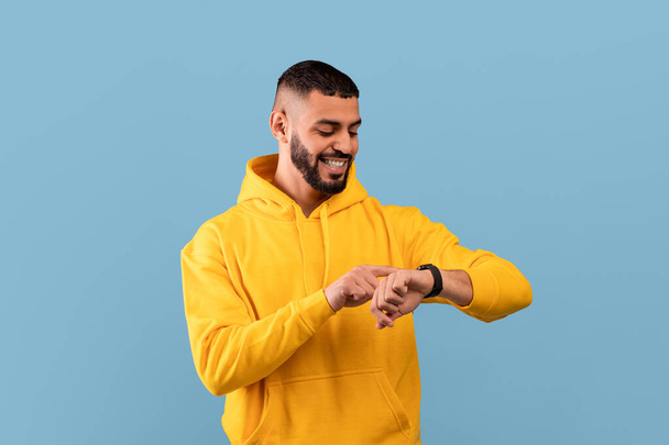 Young arab guy checking his smartwatch on blue studio background. Happy middle eastern man using fitness tracker - Фото, зображення