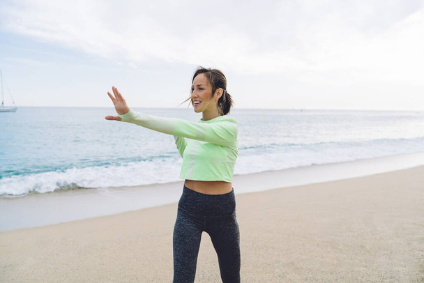 Shapely adult lady in activewear stretching arm muscles while exercising with pleasure on sandy seashore with running waves at daytime - Foto, immagini