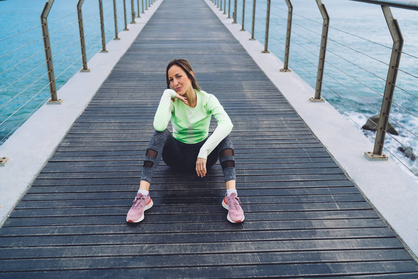 High angle of positive middle age female in sportswear and sneakers resting on wooden pier after workout and looking at camera - Foto, afbeelding