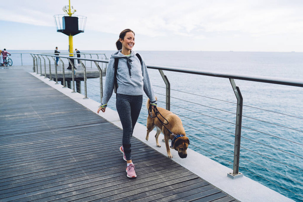 Joyful sporty young woman in casual clothes and sneakers leading on leash dog and enjoying seascape while walking on embankment on resort - Фото, зображення