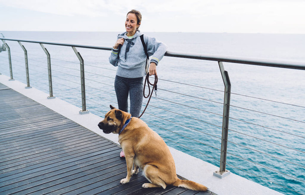 Positive young active woman standing on quay and leaning on railing while dog sitting near on leash during walk on seafront - Foto, Imagen