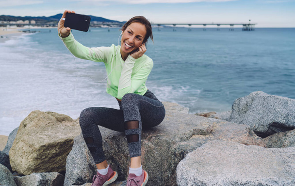 Joyful female cardio trainer in stylish tracksuit resting at coastline stones and using front mobile camera for clicking funny selfie pictures,happy fit girl in earphones shooting influence video vlog - Photo, image