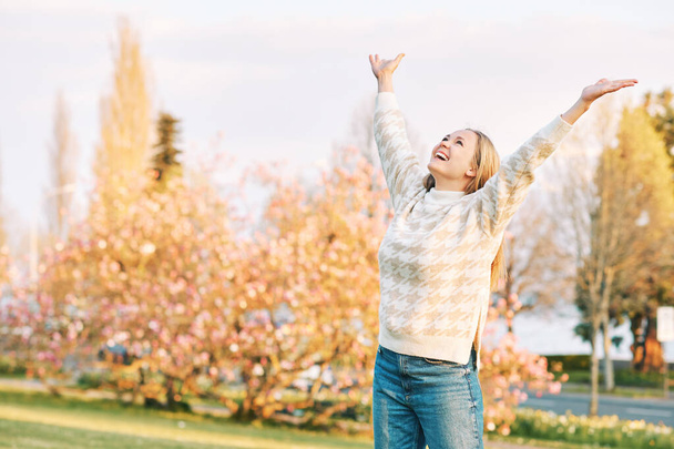 Outdoor portrait of young happy blond woman in spring park, emotional female model having fun on sunny warm day, pulling arms up wide open - Photo, Image