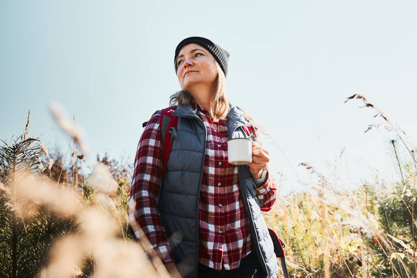 Woman taking break and relaxing with cup of coffee during summer trip. Woman standing on trail and looking away. Woman with backpack hiking through tall grass along path in mountains. Spending summer vacation close to nature - Foto, afbeelding