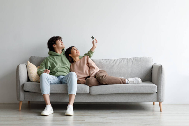 Split system. Happy young Asian couple with remote relaxing under air conditioner, sitting on couch at home, mockup - Zdjęcie, obraz