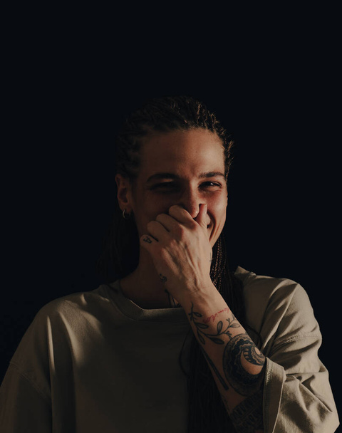 Mujer adulta con pelos trenzados y tatuajes tocándose la nariz con la mano y sonriendo. - Foto, imagen