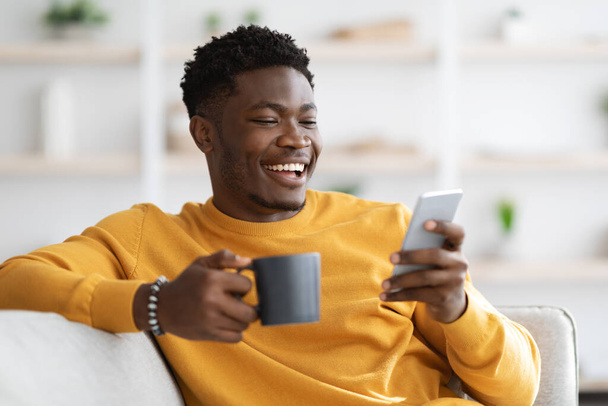 Happy african american guy reclining on couch, using smartphone - Valokuva, kuva