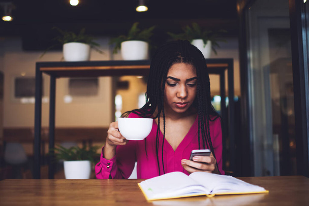Serious African American female in casual clothes of vivid color sitting at table with opened book while messaging on mobile phone and drinking coffee - Valokuva, kuva