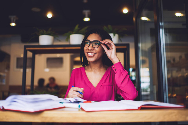 Young delighted black woman in bright casual clothes with toothy smile looking away while sitting at table in cafe with notebooks and touching eyeglasses - Foto, afbeelding