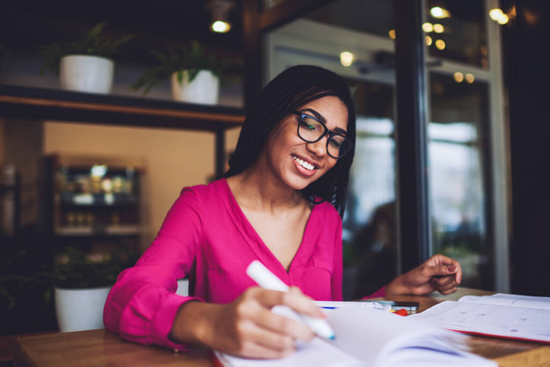 Satisfied African American woman with braids in casual clothes and spectacles looking at notebook and taking notes while preparing for exam in cafe - Foto, Imagem